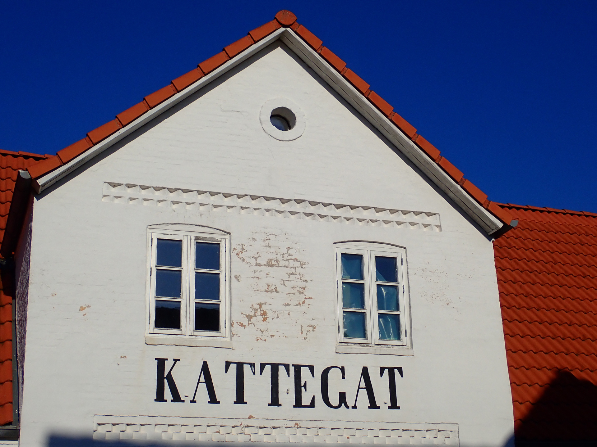 Part of the facade of a white house with a triangular roof. The word "KATTEGATT" is written on top of it.